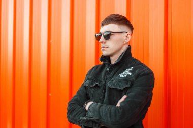 Turuncu duvarın yanında güneş gözlüklü siyah giyimleri şık bir saç modeli ile yakışıklı genç