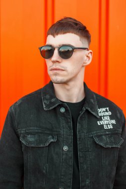 Bir saç modeli güneş gözlüğü siyah şık denim ceket sokak turuncu duvarının yakınında şık yakışıklı Amerikalı manken adam. Siyah stil