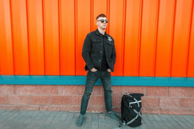 Yakışıklı genç modeli ile moda kot ceket siyah kot turuncu metal duvarın yanında bir çanta ile siyah güneş gözlüğü