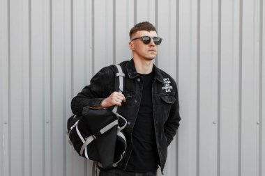 Yakışıklı genç bir siyah kot ceket içinde şık güneş gözlüğü standları sokakta gri bir duvar yakınındaki bir T-shirt ile