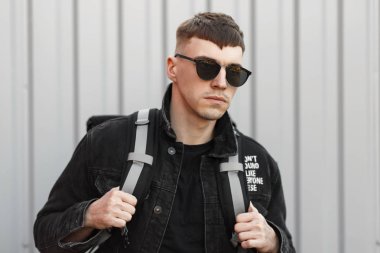 Yakışıklı genç hipster adam şık güneş gözlüğü ve bir siyah kot moda ceket gri duvarının yakınında sokakta bir çanta