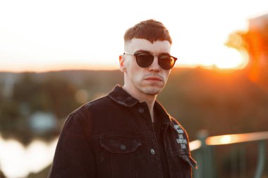 Yakışıklı genç şık Amerikan hipster adam siyah kot elbiseleri günbatımı, şık güneş gözlüğü ile