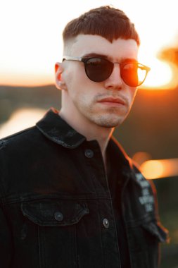 Güneşli gün batımında siyah kot pantolon giysili bir saç modeli ile gözlüklü genç bir şık adam portresi