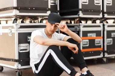 şık moda genç adam bir siyah kap ve kapsayıcılar sokakta oturan bir şerit ile siyah pantolon ile beyaz bir T-shirt