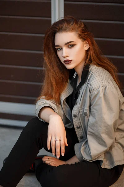时尚漂亮的年轻女子穿着时髦的夹克和黑色马球衬衫坐在门口的街道上 — 图库照片