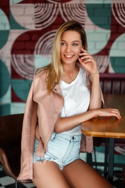 カフェのテーブルで椅子に座っているデニムのショート パンツにピンクのジャケットで笑顔で幸せな美人モデル — ストック写真