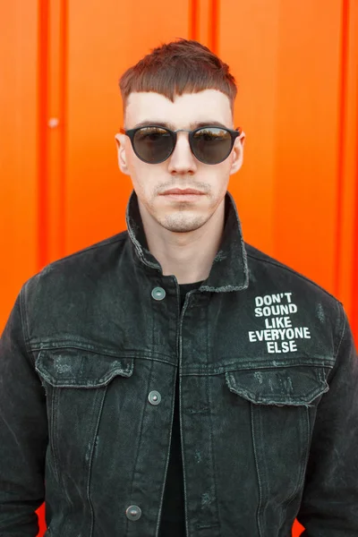 Yakışıklı genç Amerikalı adam bir turuncu duvarının yakınında siyah moda şık giysiler içinde bir saç modeli ile