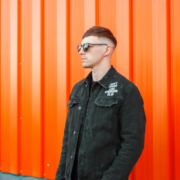 Handsome fashionable young man with hairstyle in sunglasses and a black torn jeans jacket near the orange wall