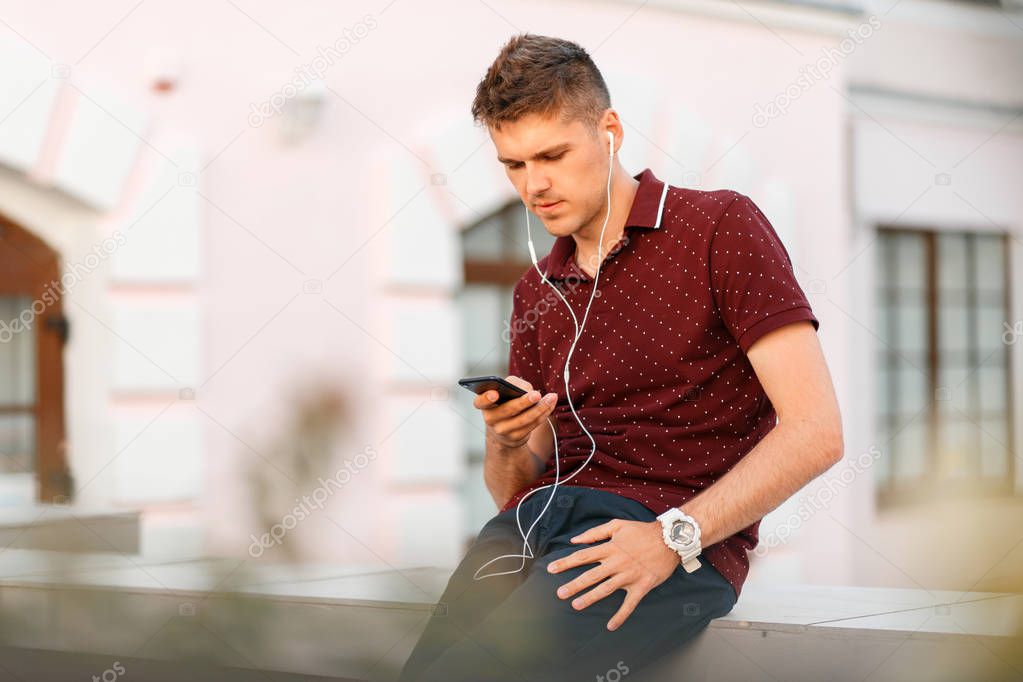 Young handsome man with phone and headphones listening to music and sitting in the city