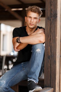 Stylish handsome young man with a haircut in a black trendy T-shirt and jeans sits on a wooden veranda