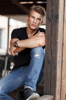 natural handsome young male model with a hairdo in a black T-shirt and jeans sits on a wooden veranda
