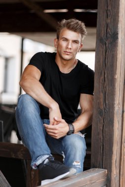 Fashion handsome young man with hair in a black shirt and jeans sitting on wooden porch
