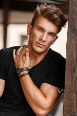Fresh portrait of a beautiful young model man in a black T-shirt on a street near a wooden pillar
