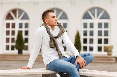 Zengin yakışıklı genç bir beyaz şık gömlek Jeans bir kazak şehirde bir bankta oturur