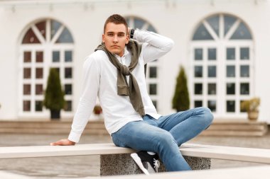 Şık yakışıklı genç başarılı adam kot Hotel dışında bir bankta oturmuş ile beyaz bir gömlek