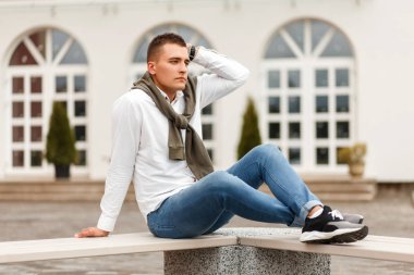 Bir kazak ile beyaz bir gömlek genç yakışıklı adam sokakta building yakınındaki bir bankta oturur