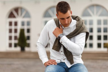 Kot pantolon ve kentin bir bankta oturmuş bir kazak ile beyaz bir gömlek de bir saat ile yakışıklı genç