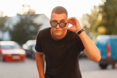 Siyah şık T-shirt moda yakışıklı genç adam güneş gözlüğü günbatımında düzeltir.