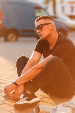 Şık hipster adam gözlüklü siyah moda giysileri günbatımında sokakta oturuyor