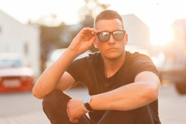 Yakışıklı hipster adam siyah moda T-shirt güneş gözlüklü bir saç modeli ile günbatımında sokakta oturuyor