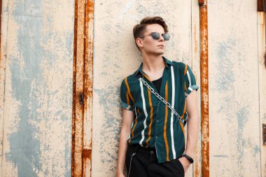 Sokakta bir metal duvar yakınındaki bir saç modeli ile bir çanta moda plaj gömlekli gözlüklü adamla yakışıklı genç modeli standları