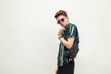 Moda güzel model adam sahilde yeşil duvarın yanında bir çanta ile renkli bir gömlek içinde güneş gözlüklü bir saç modeli ile