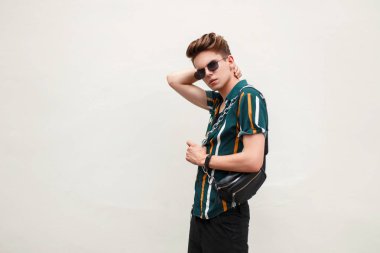 şık yakışıklı genç bir güneş gözlüğü ve çanta ayakta yeşil duvarın yanında plaj gömlekli