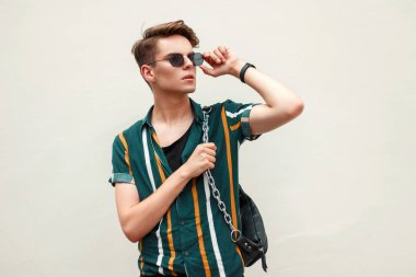 Yakışıklı genç bir şık plaj gömlekli yeşil duvar yakınındaki plajda güneş gözlüğü ayarlar