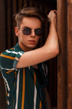 Şık yakışıklı genç model saç modeli güneş gözlüğü ve ahşap duvar yakınındaki plaj renkli gömlek ile