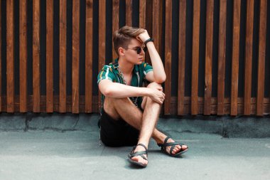 Şık genç yakışıklı bir ahşap duvar yakınında sokakta oturan şort ile şık bir gömlek