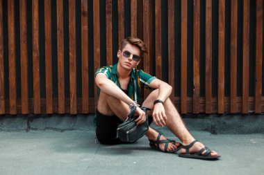 Ahşap bir duvara yakın oturan siyah şort ile plaj gömlekli gözlüklü genç moda model adam