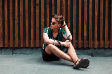 Trendy yaz giysileri ahşap bir duvara yakın oturan ayakkabı ile güneş gözlüğü ile şık yakışıklı delikanlı