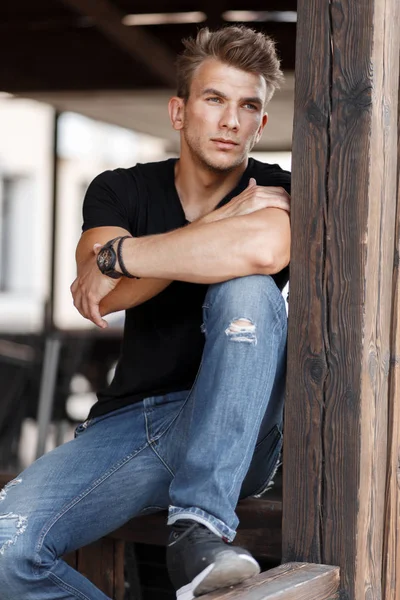 穿着时髦黑色 T恤衫和蓝色牛仔裤的英俊年轻人坐在一座木制建筑上 — 图库照片