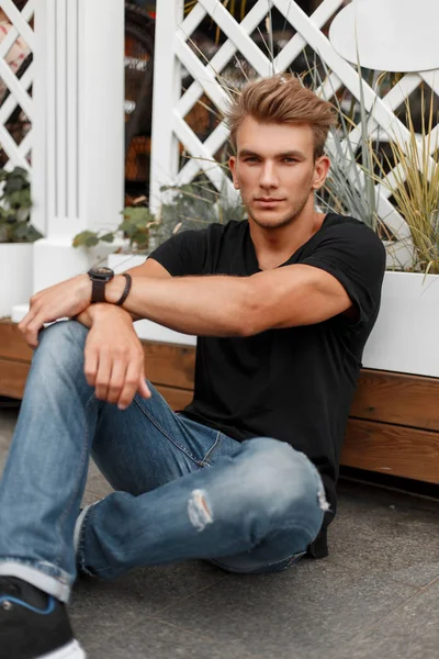 Fashionable handsome model man with a style in a black T-shirt and jeans sits near a wooden fence on the street