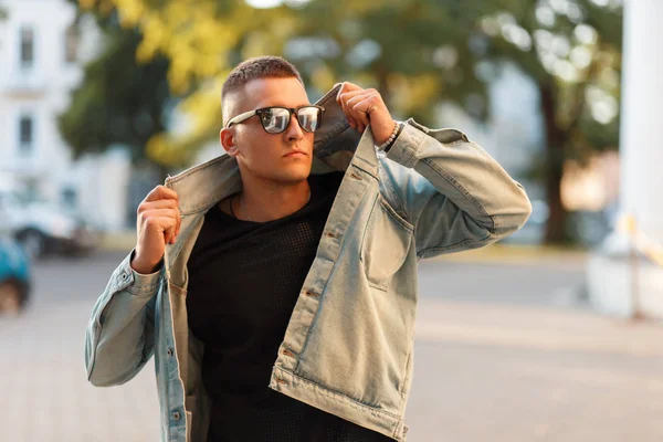 Fashionable handsome model man with a hairstyle in sunglasses and a denim jacket with a black T-shirt posing on the street