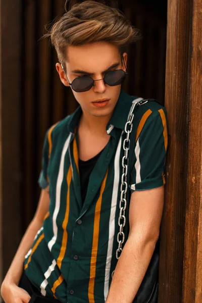 Güneş gözlüklü bir çanta ahşap duvarının yakınında şık plaj gömlekli bir saç kesimi ile bir adam yakışıklı şık genç modeli