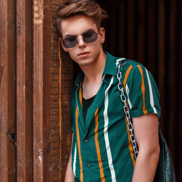Çizgili renkli gömlekli gözlüklü yakışıklı moda model adam ahşap bir duvara yakın duruyor