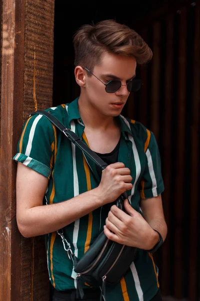 Bununla birlikte güneş gözlüklü bir plaj moda gömlek ve çanta moda yakışıklı modeli ahşap bir duvara yakın duruyor