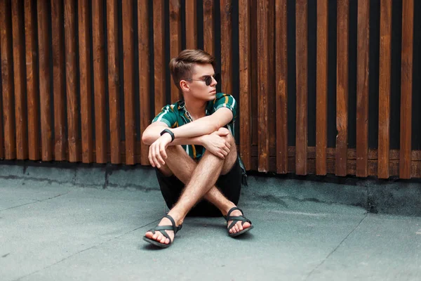 ahşap duvar yakınında sokakta oturan şort ile popüler plaj gömlekli gözlüklü şık bir saç modeli ile genç adam