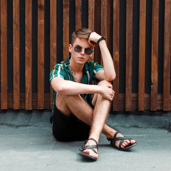 Handsome man in stylish summer clothes sits near a wooden wall on the street