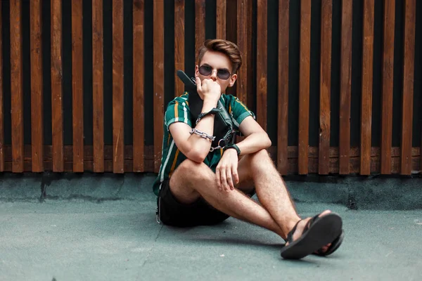 yakışıklı genç şık adam bununla birlikte güneş gözlüklü bir ahşap duvara yakın oturan plaj moda giysileri