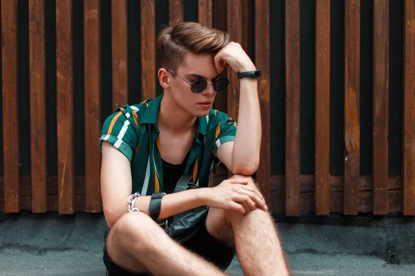 Yakışıklı model adam bununla birlikte güneş gözlüklü bir ahşap duvara yakın oturan moda giysileri