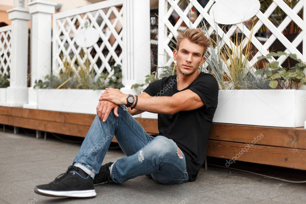 Fashionable handsome young man model with a hairstyle in a black T-shirt and jeans with sneakers sits near a wooden fence