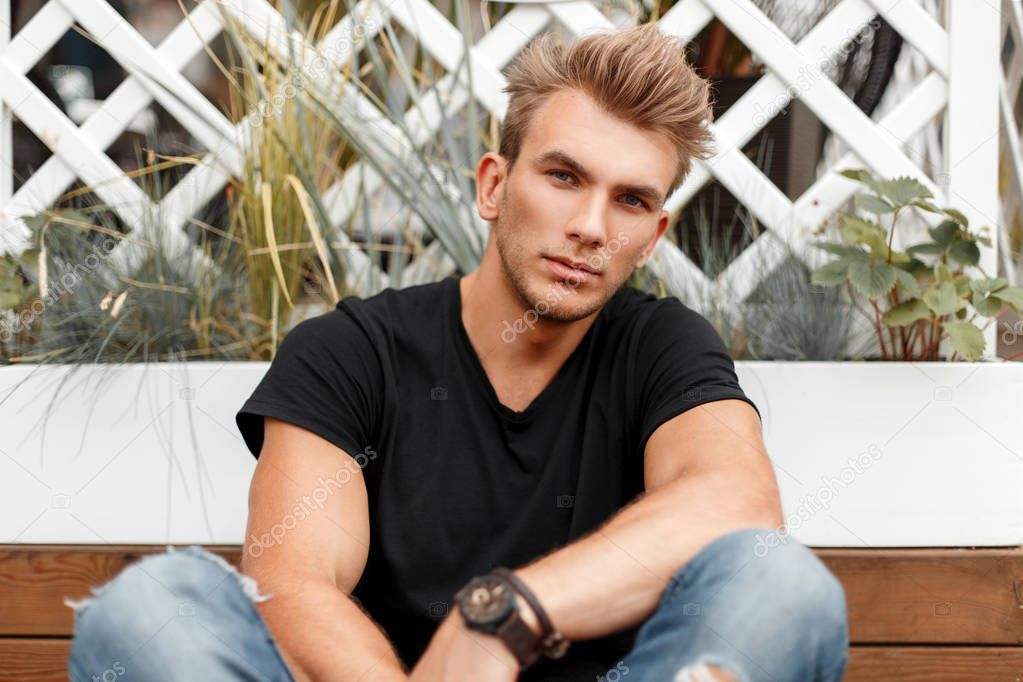 handsome fashionable guy with a hairstyle in a black T-shirt with jeans sitting near a white fence on the street