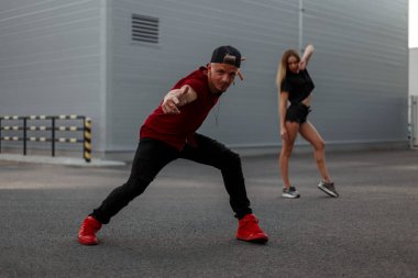 Kırmızı tişört, siyah kot pantolon ve sokakta beyzbol kap dansları ile kırmızı spor ayakkabılar şık yakışıklı genç dansçı adam. Dansçı kız arka planda