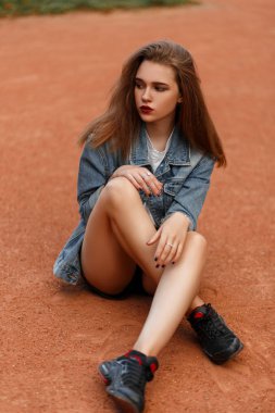 Spor ayakkabıları ile kot vintage ceket modelinde güzel genç ince kız parkta kum üzerinde oturur