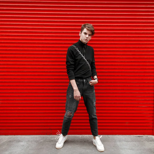 赤い金属ゲート近くに黒のプルオーバーと袋と白い靴とジーンズの若いハンサムな男が立っています ストリート スタイル — ストック写真