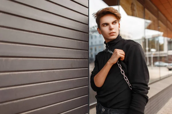 El joven guapo en la ropa a la moda negra con la bolsa posa — Foto de Stock
