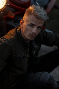 Taze bir askeri haki ceketli bir saç modeli ile yakışıklı bir moda erkek portresi bir motosiklet ışık ile akşam oturur