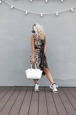Moda güzel şık genç kadın güneş gözlüğü ve gri duvarının yakınında bir desenli elbiseli şık çanta